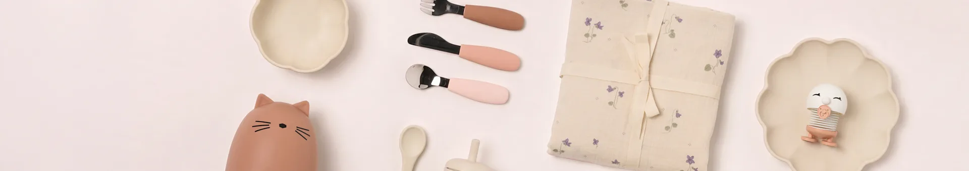 Flat lay babyservies: beige bordjes, kattenfles, roze bestek, bloemendeken, eierspeeltje.